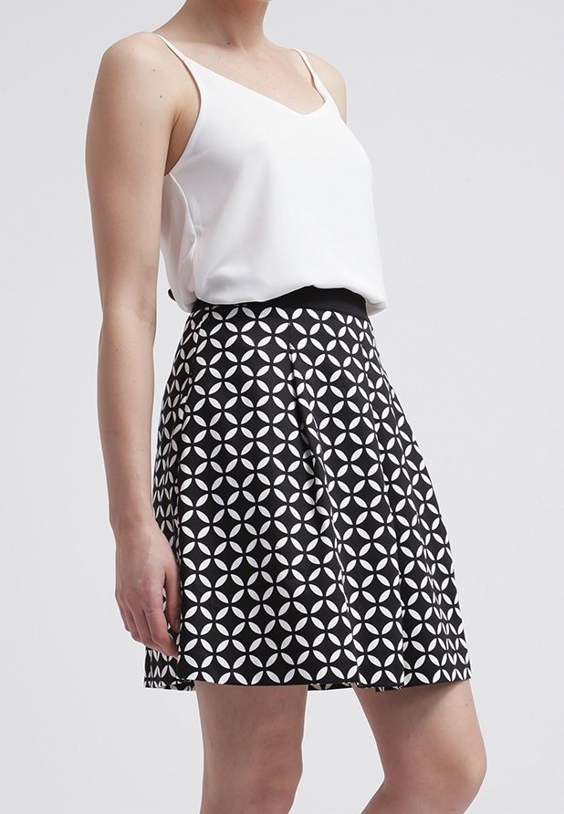 Femme portant un haut blanc sans manches et une jupe noire avec un motif géométrique de cercles blancs, debout devant un fond clair uni.