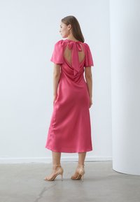 Rochie midi din satin roz cu mâneci scurte, spate deschis cu un detaliu sub formă de fundă. Textură netedă cu o siluetă fluidă, asortată cu pantofi cu toc nude.
