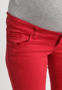 Los jeans de mezclilla rojos presentan un corte ajustado con un diseño de cinco bolsillos, un ligero desgastado y costuras visibles a lo largo de la cinturilla y los bolsillos.