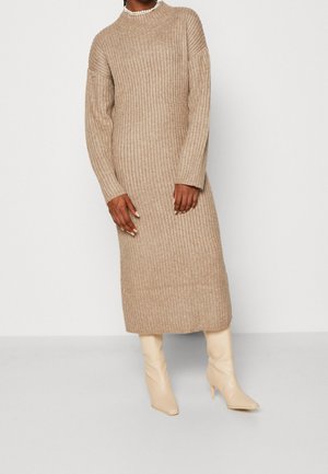 Nainen, jolla on beige, ribbineulottu, pitkähihaisen midi-mittainen neulemekko, helmikaulakoru ja kermaan sävytetyt polvenkorkuiset korkokengät, seisomassa valkoista seinää vasten.