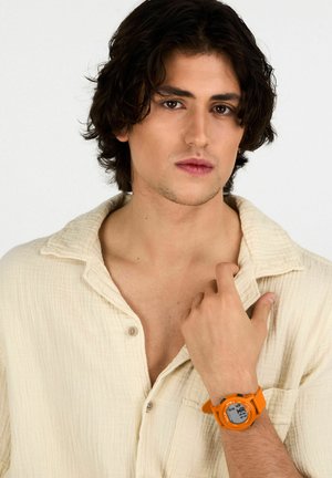 Orologio arancione con display digitale, cinturino in gomma e cassa circolare, indossato su un polso nudo; modello con camicia beige a trama.