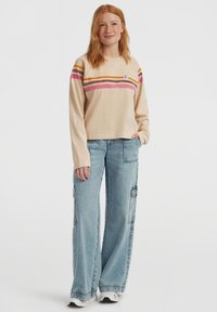 Young woman with red hair wearing a beige long-sleeve top with orange and pink stripes, light blue wide-leg jeans, and white sneakers.