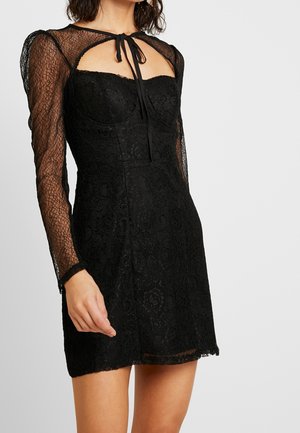 Zwarte kanten jurk met lange doorschijnende mouwen, sweetheart halslijn en lintstrik. Heeft een bloemenpatroon en een aansluitende silhouet, eindigend halverwege de dij.