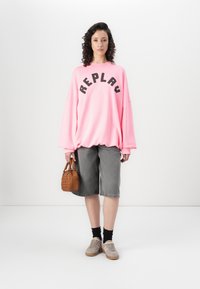 Hanorac roz oversized cu text negru "REPLAY", șorturi din denim gri, ciorapi negri, adidași bej și o geantă mică maro.