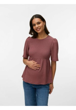 Nasmejana ženska z ramensko dolžino temnih las nosi vijoličasto teksturirano bluzo in modre kavbojke, nežno se dotika trebuha, stoji ob belem ozadju.