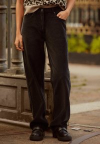Pantalons en velours côtelé noir à jambe large, avec des poches avant, une taille haute et une texture côtelée subtile. Associés à des chaussures noires.