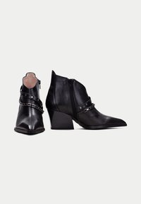Botas de cuero negro hasta el tobillo con punta afilada, acabado texturizado, cremalleras laterales y adornos de cadena plateada en la parte superior. Diseño de tacón cuadrado.