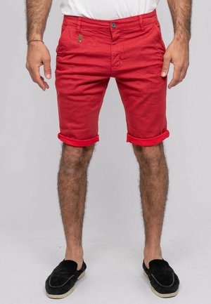 Homme portant un short rouge retroussé, une chemise blanche et des chaussures noires sans lacets, debout devant un fond uni gris clair.