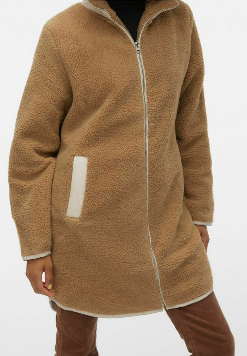 Cappotto in pile color tan con colletto alto, zip completa, dettagli color crema e tasche laterali; texture morbida e design di lunghezza intermedia.