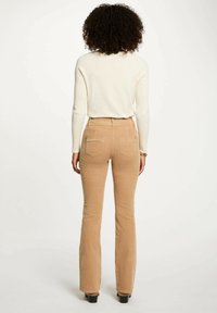 Pantaloni beige a campana con due tasche posteriori, vita alta, dettagli cuciti e una texture morbida, abbinati a un maglione aderente color crema.