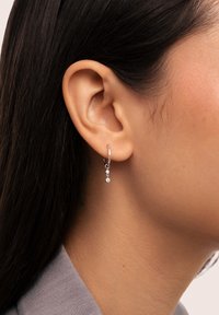 Silver hoopörhänge med två små hängande klara ädelstenar. Släta ytor, polerad finish och rundad design. Bärs på ett öra.