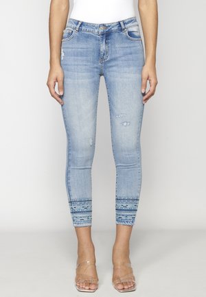 Jean slim bleu clair avec des détails déchirés et des ourlets à motifs, porté avec des talons à brides transparentes.