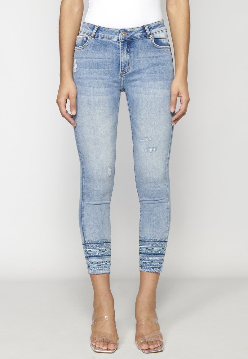 Jean slim bleu clair avec des détails déchirés et des ourlets à motifs, porté avec des talons à brides transparentes.