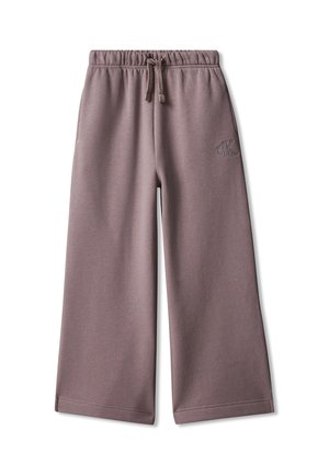 Barn med krøllet hår iført en sort denimjakke over en hvid skjorte, brede mauve bukser og sorte sko, står med hænderne i lommerne.