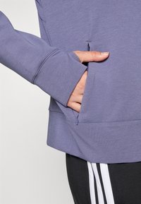 Lila sportliche Langarmoberteil mit Daumenlöchern, Reißverschlusstasche an der Seite und strukturiertem Stoff. Getragen über schwarzen Hosen mit weißen Streifen.