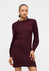 Γυναίκα με πλεξούδες, ντυμένη με μακρυμάνικο, σκούρο μοβ πλεκτό φόρεμα, που στέκεται μπροστά σε απλό φωτεινό φόντο.