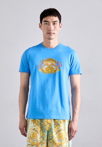 Camiseta de algodón azul con gráficos en dorado y coral que representan una cara y elementos ornamentales. Diseño de mangas cortas y cuello redondo.