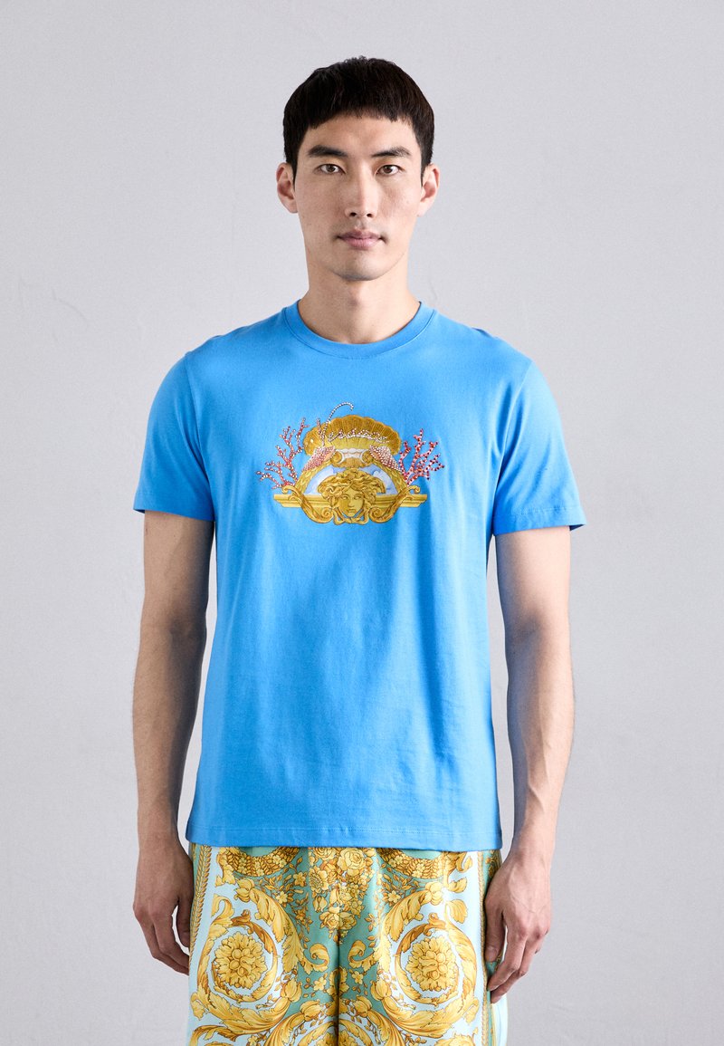T-shirt en coton bleu avec des graphiques dorés et corail représentant un visage et des éléments ornementaux. Manches courtes et design col rond.