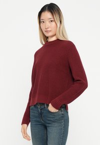 Burgundska pletena pulover z ribastim vzorcem, okroglo ovratnikom in dolgimi rokavi, v kombinaciji z denim jeansom. Pulover ima krajši kroj.