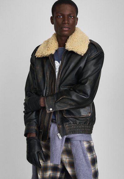 Black leather jacket with a beige shearling collar, zip closure, front pockets, and ribbed hem. Model wears black gloves and checkered pants.
