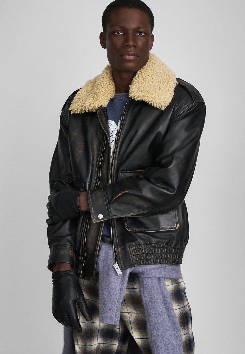 Veste en cuir noir avec un col en shearling beige, fermeture éclair, poches avant et ourlet côtelé. Le modèle porte des gants noirs et un pantalon à carreaux.