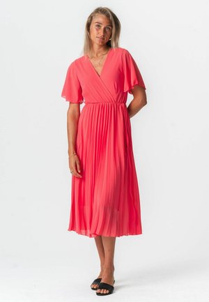 Femme portant une robe midi corail plissée avec des manches flutter et un col en V, associée à des sandales noires ouvertes en glissement, debout devant un fond blanc.
