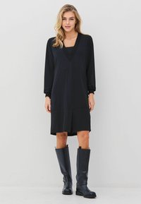 Černé šaty s dlouhým rukávem a výstřihem do V, texturovaným lemem a délkou po kolena, v kombinaci s vysokými černými botami. Hladká tkanina, pohodlný střih.