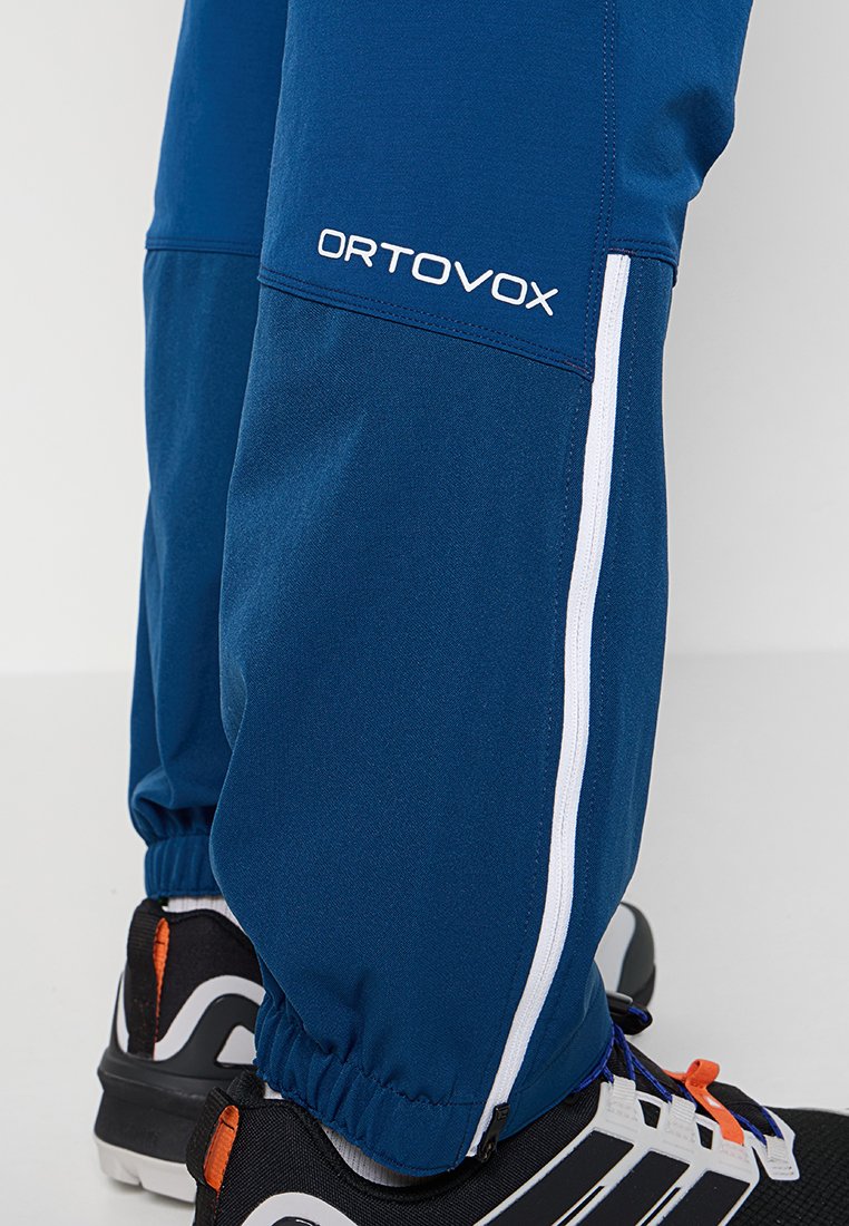 Pantalons Ortovox bleus avec des zips latéraux blancs, portés avec des chaussures de sport noires, blanches et orange sur un fond blanc.