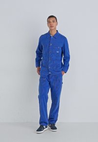 Blue denim jacket with two chest pockets, matching pants featuring cargo pockets, all in a textured fabric. Worn with dark sneakers.