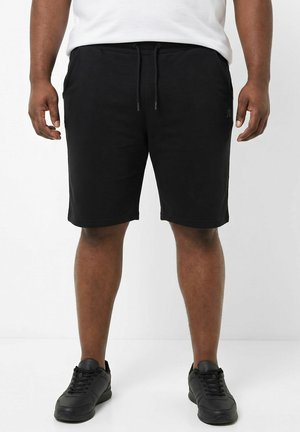 Man draagt zwarte knielange shorts met trekkoord, wit T-shirt en zwarte sneakers, staand op een effen witte achtergrond.