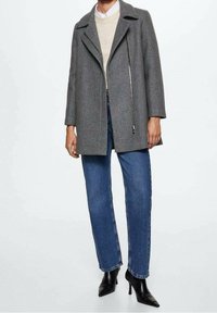 Persona vistiendo un abrigo gris con cremallera sobre un suéter beige, jeans rectos azules y botas de tacón negro con punta. 