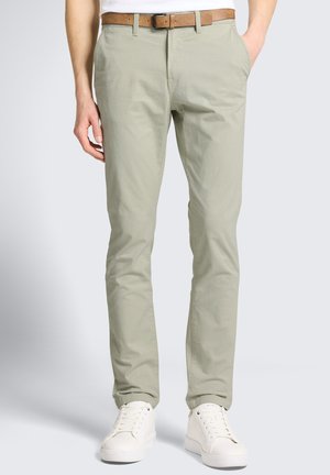 Mann trägt hellbeige Chinos, braunen Gürtel und weiße Sneaker, steht mit den Händen in den Taschen vor einem einfarbigen Hintergrund.