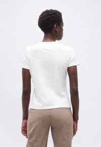 Άτομο με κοντά σγουρά μαλλιά που φοράει απλό λευκό κοντομάνικο μπλουζάκι και μπεζ παντελόνι, με την πλάτη γυρισμένη προς ένα λευκό φόντο.