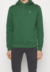 Sudadera verde de tejido suave, con un bolsillo tipo canguro y cordones ajustables. Logo rojo y blanco en el pecho, puños y dobladillo de canalé.