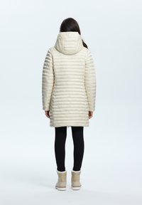 Puffy crèmekleurige jas met een capuchon, voorzien van gevilte horizontale lijnen, lange mouwen en een middellange ontwerp, gecombineerd met zwarte legging en laarzen.