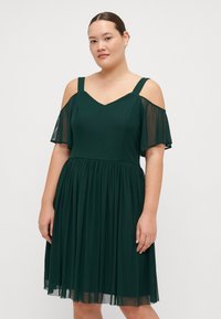 Abito verde scuro con scollatura a V, spalline scoperte e gonna fluida fino al ginocchio. Il materiale sembra morbido con maniche a farfalla trasparenti.