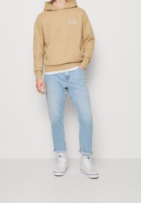 Felpa con cappuccio beige con tasca frontale e logo "CK", abbinata a jeans blu chiaro e sneakers alte bianche. Sfondo semplice.