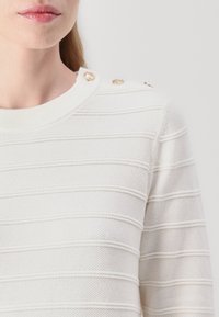 Hvit strikket genser med horisontale striper, ribbet halslinning, og gullknapper på skulderen. Teksturen ser myk ut med en lett vev.