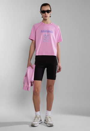 Roze cropped t-shirt met grafische tekst, gecombineerd met zwarte fietsbroek. Model houdt een bijpassende roze jas vast; draagt zwarte zonnebril en witte sneakers.