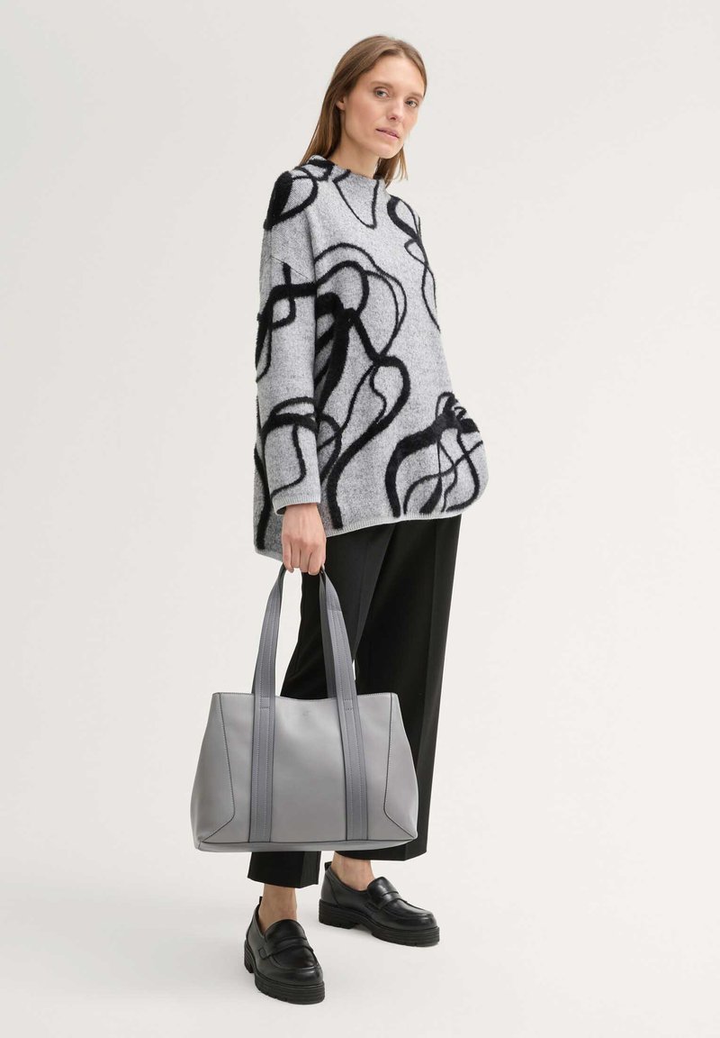 Femme portant un pull gris avec des lignes noires abstraites, un pantalon noir raccourci, des mocassins noirs, tenant un grand sac fourre-tout gris.