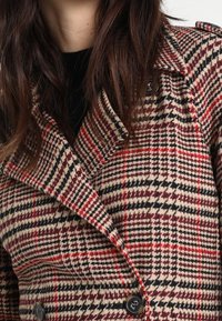 Cappotto doppiopetto con motivo a soffici in beige, nero e rosso. Presenta grandi bottoni neri e un colletto strutturato. Tessuto testurizzato.