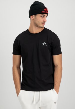 Camiseta de algodón negra con mangas cortas, que presenta un pequeño logo blanco en el lado izquierdo del pecho y un diseño de cuello redondo. El modelo lleva pantalones blancos.