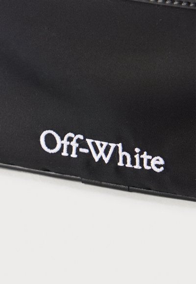 Μαύρη υφασμάτινη τσάντα με λευκό κεντημένο λογότυπο «Off-White» κοντά στην κάτω άκρη σε φωτεινό φόντο επιφάνειας.