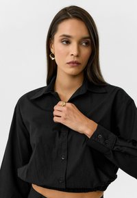 Zwarte cropped button-up shirt met een kraag, lange mouwen en manchetten. Heeft een gathers zoom en is gecombineerd met een gouden ring en oorbellen.