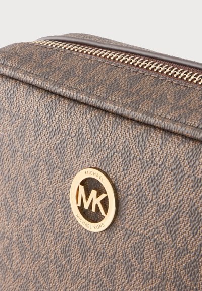 Καφέ υφασμάτινη τσάντα Michael Kors με χρυσλό λογότυπο MK και λεπτομέρεια επάνω φερμουάρ σε ανοιχτό φόντο.