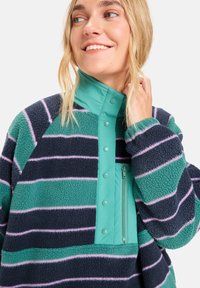 Fleece pulover s prugama u tamnoplavoj, tirkiznoj i lavandi. Ima visoki ovratnik s kopčama na dugmad i džep s patentnim zatvaračem sprijeda.