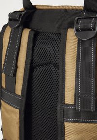 Tan en zwarte rugzak met mesh padding, voorzien van verstelbare schouderbanden en duurzaam materiaal. Gedetailleerde stiksels zichtbaar op de bandjes en randen.