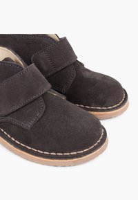 Scarpe in suede nere con punta arrotondata e cinturino in velcro. Cuciture beige evidenti lungo la suola e un interno liscio.