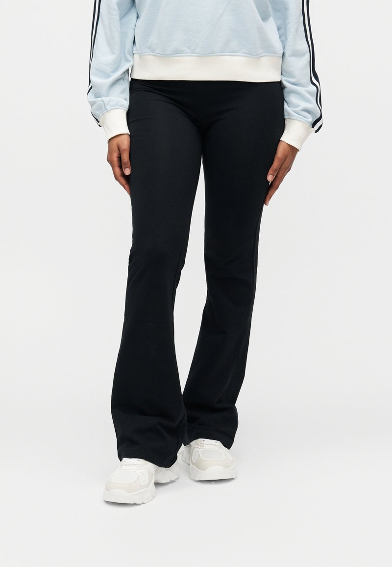 Personne portant un pantalon noir évasé, des baskets blanches et un sweat-shirt bleu clair avec des rayures noires sur les manches, sur un fond blanc.