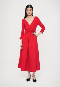 Piros, hosszú ujjú, átlapolt ruha V-nyakkivágással és csomózott derékkal. Laza szoknyával és sima textúrával rendelkezik. Fekete magassarkúval viselve.